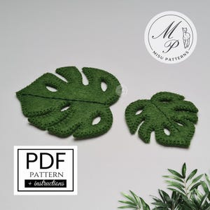 Könnte beinhalten: Zwei grüne Monstera-Blätter aus Filz, eines groß und eines klein, mit ausgeschnittenen Löchern, werden auf einer weißen Oberfläche gezeigt. Die Blätter haben genähte Ränder. Ein weißes Quadrat mit dem Text "PDF PATTERN + instructions" befindet sich unten links.