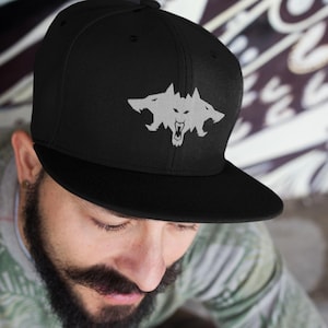 May include: Black baseball cap with a flat brim. The cap features a white graphic of a three-headed wolf. The cap is worn by a person with a beard and mustache.