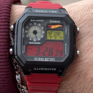 May include: Black and red Casio digital watch with a rectangular face. The watch features a red band and a digital display showing the time. The watch face includes the text "CASIO", "WORLD TIME", and "10 YEAR BATTERY".