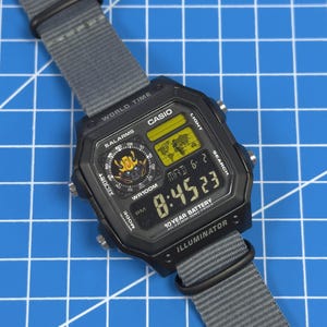 May include: Black Casio digital watch with a gray nylon strap. The watch face displays the time, date, and other functions. The watch has a rectangular shape and features the text "CASIO" and "ILLUMINATOR". The strap is secured with a black buckle.