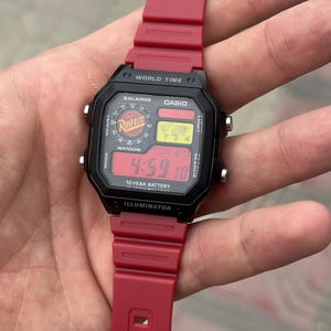 Pode incluir: Um relógio digital Casio World Time com caixa retangular preta e pulseira de resina vermelha. O mostrador exibe a hora 4:59, um mapa-múndi e o logotipo Rust-eze. O texto inclui World Time, 10 Year Battery e a palavra Illuminator.