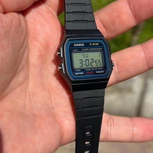 May include: Black Casio F-91W digital watch. The watch face displays the time, day, and date. The watch has a black resin band and a blue border around the digital display. The watch is water resistant.