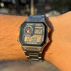 May include: A silver Casio digital watch with a black bezel is displayed on a wrist. The watch face shows the time, date, and world time features. The watch has a stainless steel band and features the text "WORLD TIME", "CASIO", and "ILLUMINATOR".
