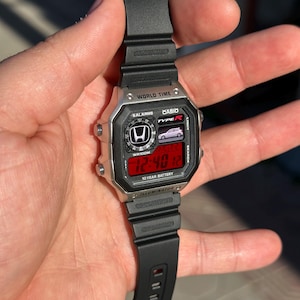 May include: A silver and black digital watch with a black band. The watch face displays the time in red digits and features the Honda logo and the text "TYPE R". The watch also has the text "WORLD TIME" and "CASIO".