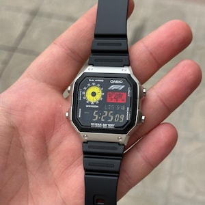 May include: A silver Casio digital watch with a black band. The watch face displays the time, date, and a yellow and black graphic. The watch has the text "CASIO" and "FI" on the face. The watch band is made of black rubber.
