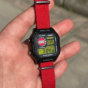 May include: A black Casio digital watch with a red nylon strap. The watch face displays the time and features the Lego logo. The watch is water resistant and has a 10-year battery.