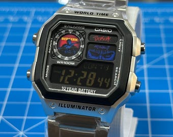 Könnte beinhalten: Eine silberfarbene Digitalarmbanduhr mit Metallgliederarmband. Das Zifferblatt hat einen schwarzen Rand und die Aufschriften "WORLD TIME", "CASIO", "BERSERK" und "ILLUMINATOR". Das Display zeigt "12:28 44" und eine kreisförmige Grafik mit dem Wort "BERSERK".