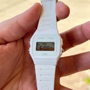 Könnte beinhalten: Eine weiße Casio F-91W Digitaluhr mit weißem Armband. Das Zifferblatt zeigt die Uhrzeit, den Tag und das Datum an. Die Uhr wird in einer Hand gehalten.
