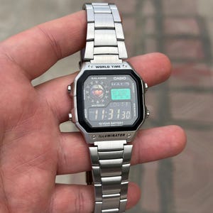 Könnte beinhalten: Eine silberne digitale Casio-Uhr mit einem schwarzen quadratischen Zifferblatt. Die Uhr zeigt den Text "WORLD TIME", "CASIO" und "HALO" an. Das Uhrenarmband besteht aus silbernen Metallgliedern. Die angezeigte Uhrzeit ist 11:31:30.