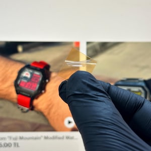 Puede incluir: Un pequeño trozo de plástico transparente, de forma cuadrada, se sostiene en una mano enguantada. El plástico parece estar ligeramente teñido de amarillo. En el fondo, se ve un reloj digital con correa roja.