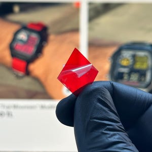 Puede incluir: Un pequeño cuadrado rojo transparente con un respaldo despegable se sostiene en una mano enguantada. El cuadrado está ligeramente inclinado, revelando su forma. En el fondo, se ve un reloj con una correa roja y una pantalla digital.