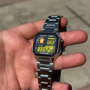May include: A silver-tone digital wristwatch with a metallic link band. The rectangular face features a black border, a yellow digital display showing 11:52:58, a world map graphic, and a small circular icon of a yellow character. Text includes CASIO, WORLD TIME, WR100M, and 10 YEAR BATTERY.