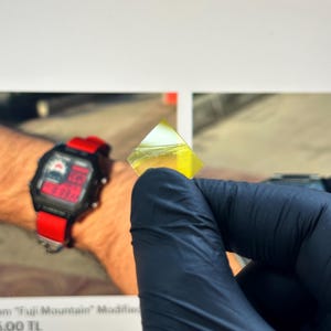 Puede incluir: Un pequeño objeto geométrico transparente de color amarillo verdoso es sostenido por una mano enguantada. El objeto está enfocado, con un fondo borroso que incluye un reloj digital con una correa roja.