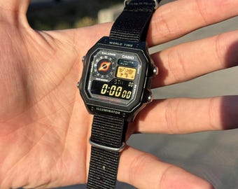 May include: Black digital watch with a black nylon strap. The watch face displays the time and world time features. The watch has the text "WORLD TIME" and "CASIO" printed on the face.