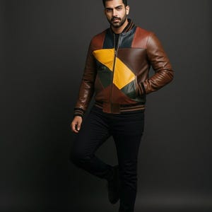 Può includere: Giacca in pelle marrone con un design patchwork con pannelli gialli, verdi e marroni. La giacca ha colletto e polsini a coste e chiusura con cerniera. Il modello indossa pantaloni e scarpe neri.