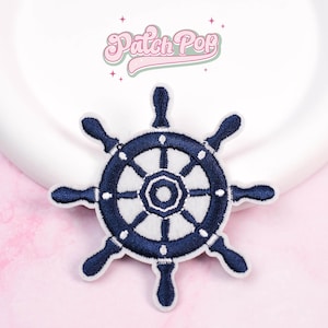 May include: Embroidered navy blue ship's wheel patch with white accents. The patch features a detailed design of a nautical steering wheel, perfect for adding a maritime touch to clothing or accessories. The text "Patch Pop" is above the patch.
