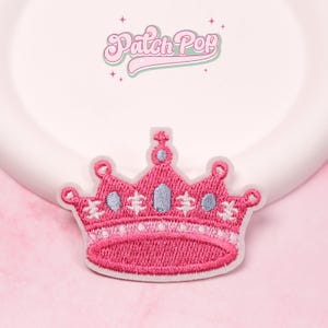 Pode incluir: Um patch de coroa bordada rosa com detalhes em azul claro e branco. O patch tem uma borda branca e está sobre um fundo rosa. O texto "Patch Pop" está acima da coroa.