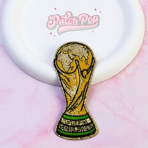 May include: A gold glitter embroidered patch shaped like the FIFA World Cup trophy, featuring a globe at the top and the text FIFA WORLD CUP in black on a green base. The patch is displayed below the pink text Patch Pop on a white circular background.