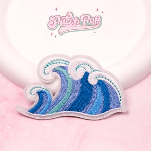 以下が含まれることがあります： 青と白の色合いの波の刺繍パッチ。パッチは、明るい灰色の境界線を持つ詳細なデザインが特徴です。「Patch Pop」というテキストが波の上に筆記体で表示されています。