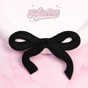 Könnte beinhalten: Ein schwarzer, gestickter Schleifenaufnäher mit detailliertem Design. Der Aufnäher ist auf einer weißen Oberfläche mit rosa Hintergrund zentriert. Der Text "Patch Pop" ist oben in der Mitte sichtbar.