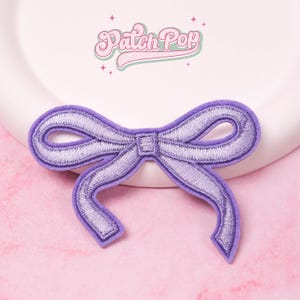 Op de afbeelding: Een lavendelkleurige geborduurde strikpatch met een fluwelen textuur. De strik heeft een gedetailleerd gestikt ontwerp en is geplaatst tegen een roze achtergrond. De tekst "Patch Pop" is zichtbaar in het bovenste deel van de afbeelding.