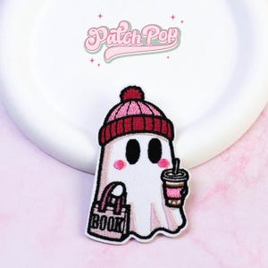 Può includere: Una toppa ricamata raffigurante un fantasma bianco che indossa un berretto rosa e rosso scuro. Il fantasma ha occhi neri, guance rosa e tiene in mano una tazza di caffè marrone e una borsa nera con la parola BOOK stampata in bianco.