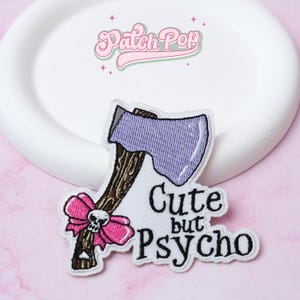 Puede incluir: Parche bordado con un hacha morada con mango marrón y un lazo rosa con una calavera. El texto "Cute but Psycho" está debajo del hacha. El parche está sobre una superficie blanca con el texto "Patch Pop" en rosa.
