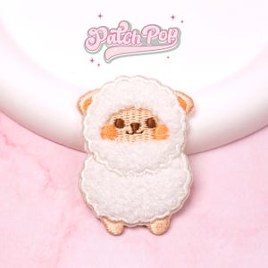 Puede incluir: Parche bordado con un diseño de oveja con un cuerpo blanco y esponjoso, orejas marrones y mejillas rosadas. El parche tiene un borde cosido y el texto "Patch Pop" en rosa, en cursiva, encima de la oveja.