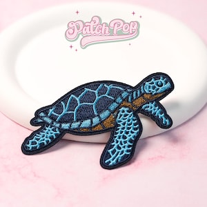Può includere: Toppa ricamata di una tartaruga marina blu e marrone. La tartaruga ha un guscio blu scuro con un motivo geometrico azzurro. Il testo "Patch Pop" è sopra la tartaruga in rosa.
