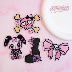 May include: A collection of embroidered patches featuring a pink skull with crossbones, a bunny, a black boot with a purple rose, and a pink bow with a skull. The patches have a black and pink color scheme.