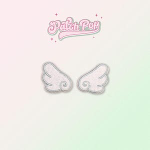 Könnte beinhalten: Zwei weiße, gestickte Bügelbilder in Form von Engelsflügeln. Die Flügel sind mit silbernem Garn umrandet.