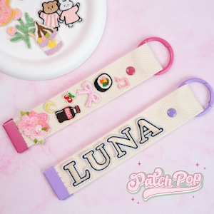 May include: Two cream-coloured keychains with colourful embroidered patches. One keychain has the name "LUNA" embroidered in blue. The other keychain has patches of a cherry, sushi, and a pink bow. Both have pink and purple accents.