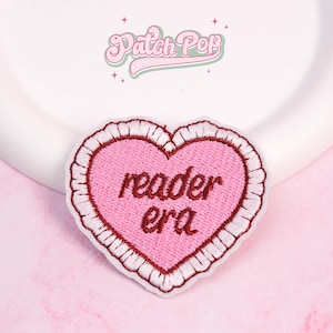 Puede incluir: Parche rosa en forma de corazón con las palabras "reader era" bordadas. Tiene un borde blanco con volantes y un fondo rosa claro. El logotipo "Patch Pop" es visible en la parte superior. Ideal para ropa o accesorios.
