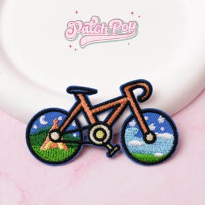 May include: Embroidered bicycle patch with an orange frame and blue wheels. The wheels depict landscape scenes: one with a tent and moon, the other with clouds and sky. The patch has a black border and pink text: "Patch Pop".