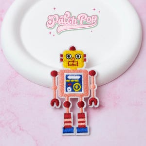 Puede incluir: Parche bordado de robot con cabeza amarilla, cuerpo rosa y detalles rojos y azules. El robot tiene un cuerpo cuadrado con un diseño similar a una pantalla. El parche está sobre un plato blanco con el texto "Patch Pop" en rosa.