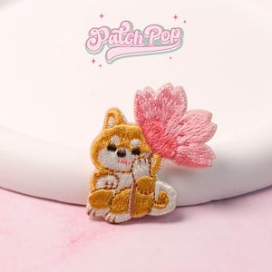 May include: Embroidered patch featuring a Shiba Inu dog holding a pink flower. The dog is in shades of brown, white, and gold. The patch has the text "Patch Pop" in a pink, cursive font.