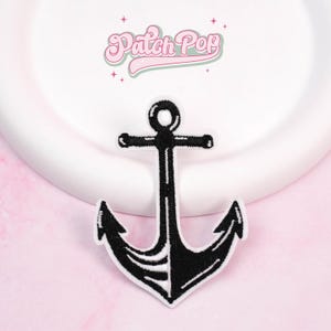 May include: Black and white embroidered anchor patch. The anchor design is centered on a white background, with the text "Patch Pop" in pink script above. The patch is on a pink surface.