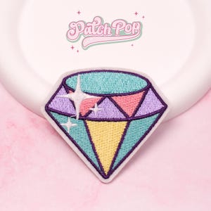 May include: Embroidered diamond patch with a colorful design. The patch features a light blue, purple, pink, and yellow color scheme, with a white starburst design. The patch is set against a pink background and a white plate. The text "Patch Pop" is at the top.