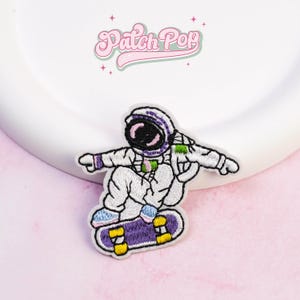 May include: Embroidered patch featuring an astronaut skateboarding. The astronaut is wearing a white spacesuit with purple and green accents. The skateboard is purple with yellow wheels. The patch has the text "Patch Pop" in a pink, cursive font.