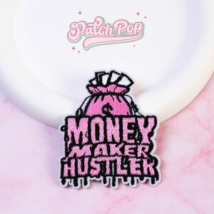 May include: An embroidered patch featuring a pink money bag with white cash sticking out at the top. Below the bag, the text reads MONEY MAKER HUSTLER in bold pink letters with a black outline and a dripping effect at the bottom edge.