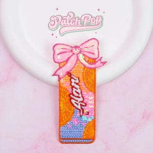 May include: A rectangular sequined patch with a pink bow at the top. The patch is orange with the word "Alan" in white script. The bottom of the patch is blue with a floral design. The patch is on a white plate and pink background.