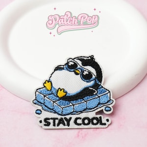 Puede incluir: Parche bordado con un pingüino de dibujos animados con gafas de sol, tumbado en un bloque de hielo azul. El parche tiene el texto "STAY COOL" en blanco. El parche está sobre un plato blanco.