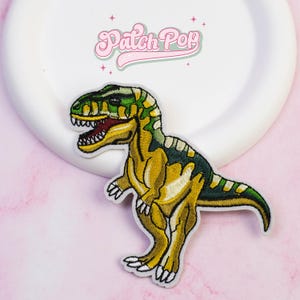 Op de afbeelding: Geborduurde patch met een Tyrannosaurus Rex dinosaurus in geel, groen en bruin. De patch heeft een gedetailleerd ontwerp met zichtbare tanden en klauwen. De tekst "Patch Pop" staat in roze, cursieve letters.