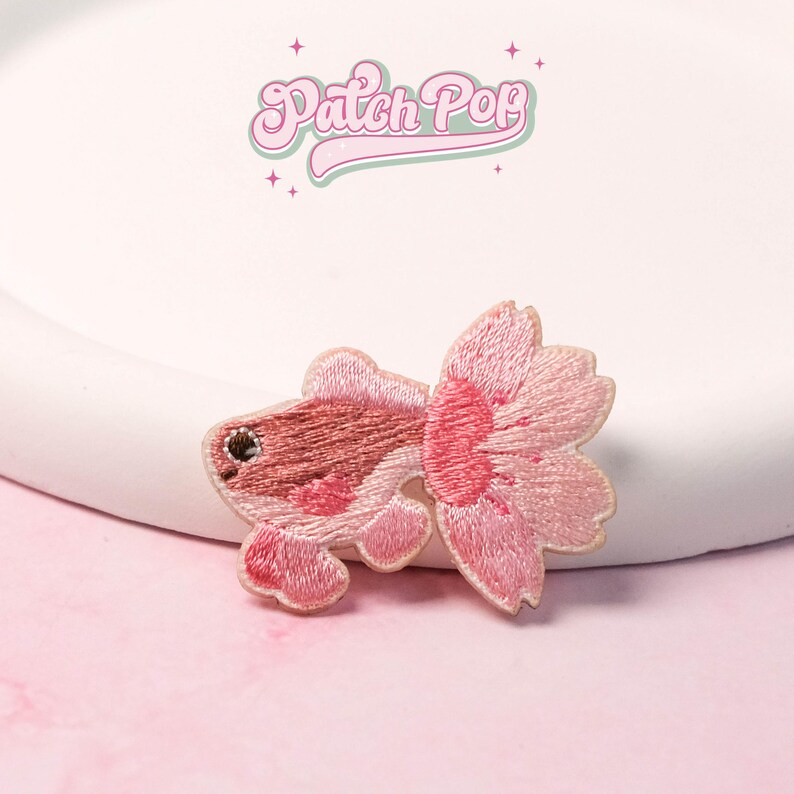 May include: Embroidered patch of a pink goldfish with a large, flowing tail. The fish has a detailed design with varying shades of pink and a small, dark eye. The patch is set against a white background with the text "Patch Pop" in a cursive font.