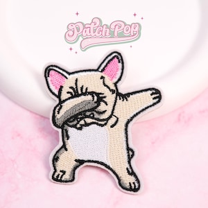 May include: Embroidered patch of a tan French Bulldog in a dabbing pose. The dog has pink inner ears and a black nose and mouth. The patch has a white border and the text "Patch Pop" in pink and green.