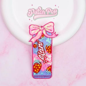 May include: A sequined patch featuring a pink bow and a rectangular design with the word "Alani" in white script. The design includes red strawberries and a blue background. The patch is likely for clothing or accessories.