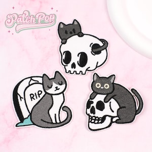 Può includere: Tre toppe ricamate a tema gatto spettrale. Una toppa mostra un gatto nero su un teschio, un'altra mostra un gatto su una lapide con "RIP" e la terza mostra un gatto su un teschio. Le toppe hanno uno sfondo bianco e dettagli neri e grigi.