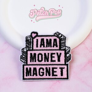 Może przedstawiać: Haftowana naszywka przedstawiająca trzy ułożone w stos różowe prostokątne bloki z czarnym napisem I AM A MONEY MAGNET. Wzór zawiera małe różowe serce na górnym bloku oraz stosy szaro-białych banknotów po obu stronach.
