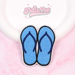 Puede incluir: Parche bordado con un par de chanclas azules con contornos negros. El parche tiene un borde blanco y está sobre un fondo blanco. El texto "Patch Pop" se muestra en rosa, en fuente cursiva.