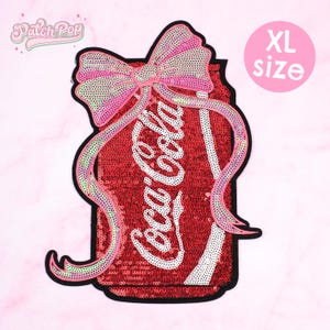 Puede incluir: Parche XL con lentejuelas en forma de lata de Coca-Cola con un lazo rosa. La lata es roja con texto blanco. El lazo y la cinta son rosas, verdes y blancos. El parche tiene un borde negro.